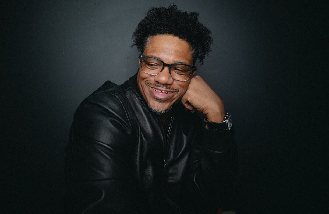 A black man with a confident smile, wearing glasses and a black leather jacket, rests his head on his hand against a dark background. The photo exudes a relaxed yet self-assured vibe, lifestyle image blending professionalism with warmth.